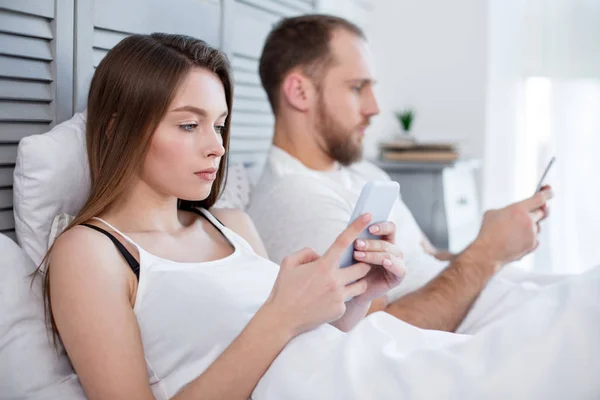 O femeie serioasă și un bărbat întins în pat cu telefoanele lor — Fotografie, imagine de stoc