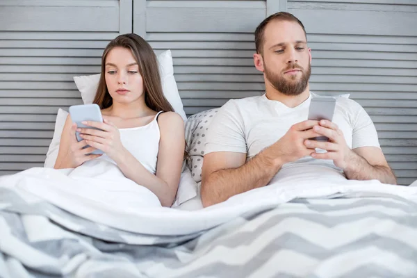 O femeie hotărâtă și un bărbat întins în pat cu telefoanele lor — Fotografie, imagine de stoc