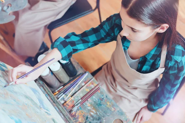 Chestnut haired girl sitting at easel while painting — Stock Photo, Image