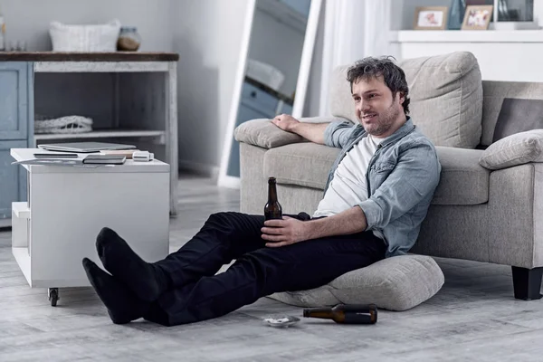 Улыбающийся неопрятный мужчина пьет алкоголь и смотрит телевизор дома. — стоковое фото