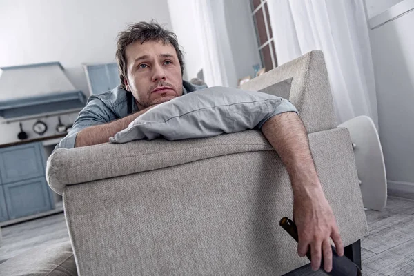 Сумний чоловік тримає пляшку алкоголю, розслабляючись на дивані — стокове фото