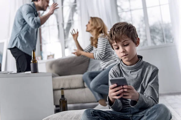 Tranquilo niño sosteniendo un teléfono inteligente mientras sus padres tienen una fuerte pelea —  Fotos de Stock