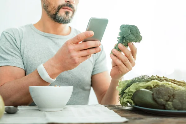 Hombre concentrado ocupado sosteniendo un brócoli usando su teléfono celular . — Foto de Stock