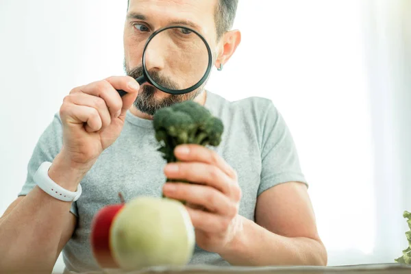 Homem curioso procurando segurando um brócolis olhando através de lupa . — Fotografia de Stock