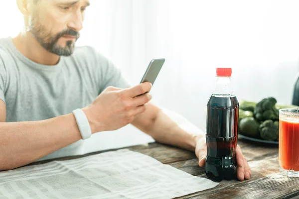 Hombre concentrado e inquebrantable usando un celular tocando la botella . — Foto de Stock