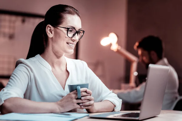 Nöjd glasögonprydd kvinna håller en kopp och använder den bärbara datorn. — Stockfoto