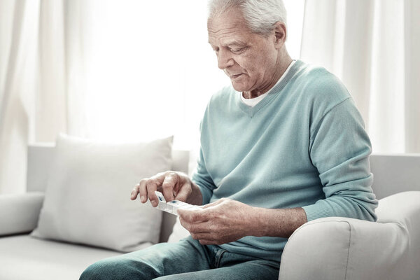 Aged pleasant man taking a pill and looking at tablets.