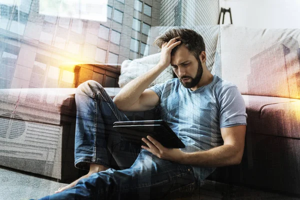 Эмоциональный человек хмурясь, сидя на полу со своим планшетом — стоковое фото