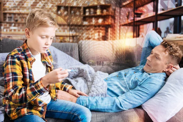 Chico asombrado mirando el termómetro mientras estaba sentado con su padre enfermo — Foto de Stock