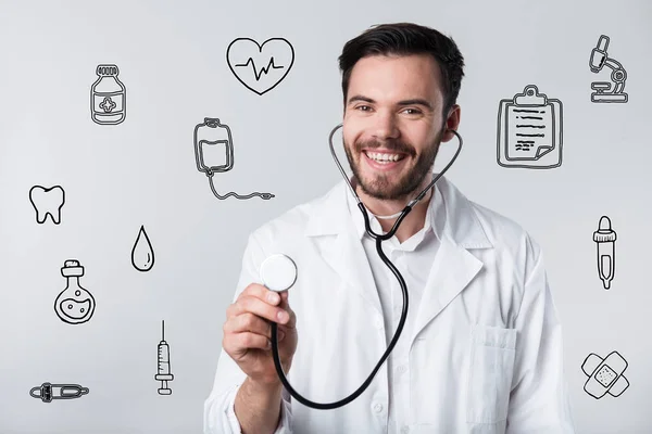 Médico barbudo positivo sonriendo y sosteniendo su estetoscopio — Foto de Stock