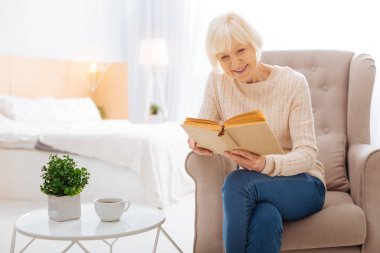 Sevimli gülümseyen yaşlı kadın bir koltukta otururken bir kitap okuma