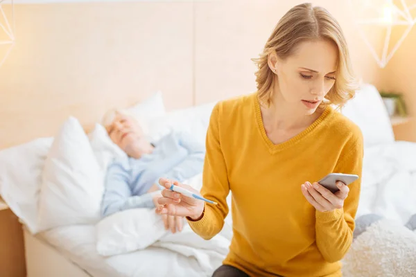 Serieuze jonge vrouw probeert te bellen met een arts terwijl een thermometer — Stockfoto