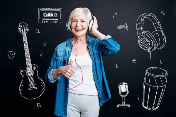 Mujer mayor positiva poniendo su pulgar hacia arriba mientras escucha música —  Fotos de Stock