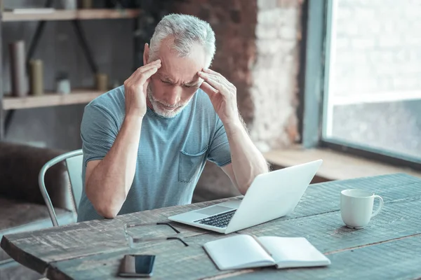 Ill uitgeput man zijn hoofd aan te raken en zitten bij de tabel. — Stockfoto