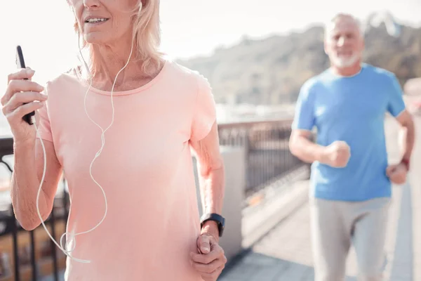 Mujer anciana concentrada corriendo y sosteniendo el teléfono inteligente . — Foto de Stock