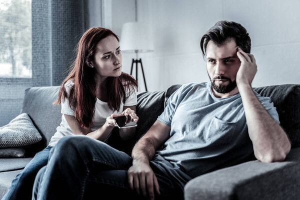 Serious redhead woman holding cellphone and looking at her husband.