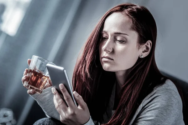 Upprörd rödhårig kvinna håller ett glas och med hjälp av mobiltelefon. — Stockfoto