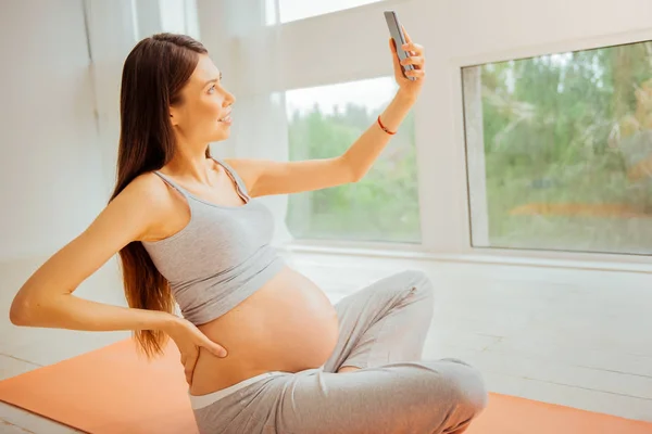 Elégedett terhes nő ül a földön és a gazdaság smartphone. — Stock Fotó
