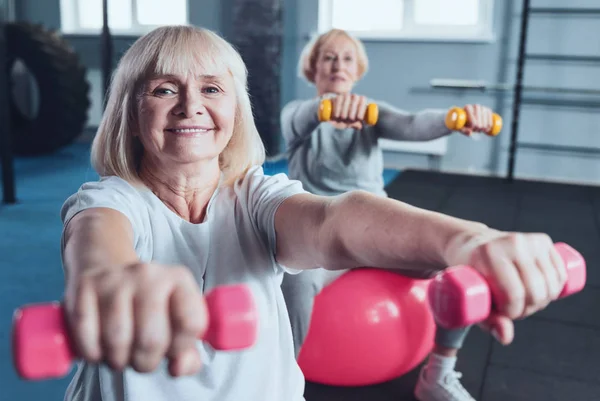 Encantadora anciana sonriendo para la cámara mientras levanta pesas — Foto de Stock