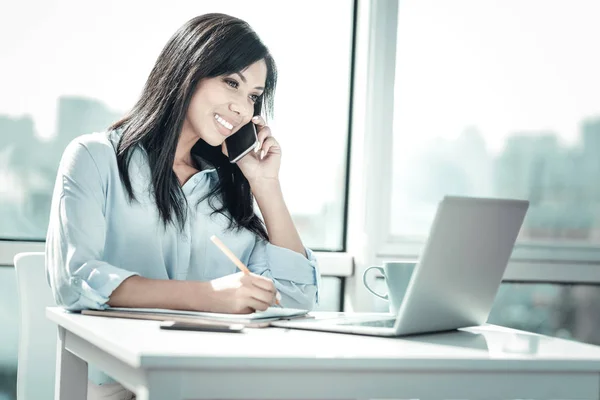 Occupied smiling woman having phone conversation and writing. — Stock Photo, Image