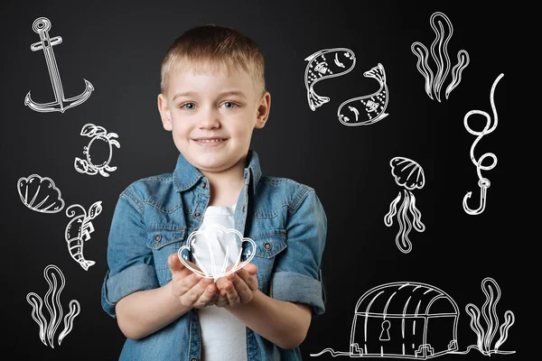 Kleiner glücklicher Junge lächelt, während er eine schöne Muschel in der Hand hält — Stockfoto
