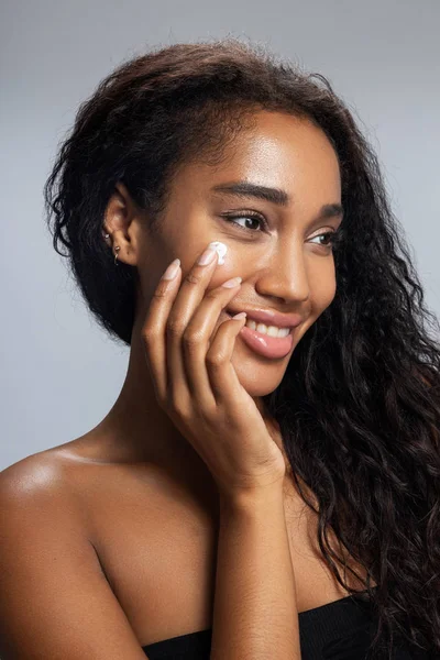 Sonriendo mujer afroamericana está aplicando crema facial — Foto de Stock