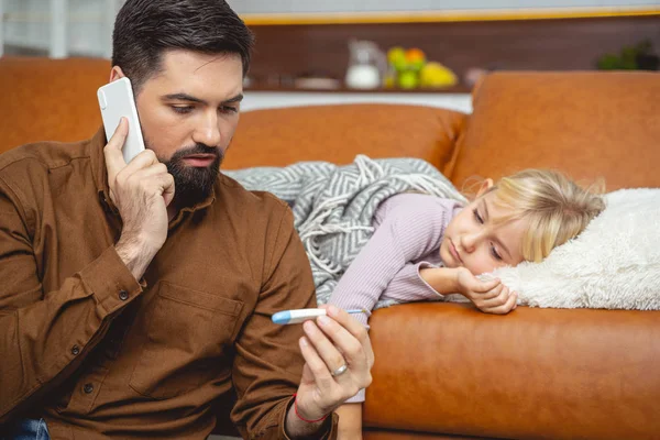 Bearded man taking daughter temperature and calling doctor — Stock Photo, Image