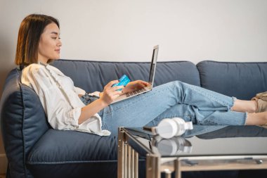 Asyalı iş kadını kanepede uzanıyor ve internetten ödeme yapıyor.