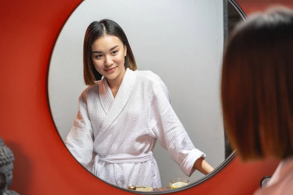 Молодая милая азиатка смотрит в зеркало в помещении — стоковое фото