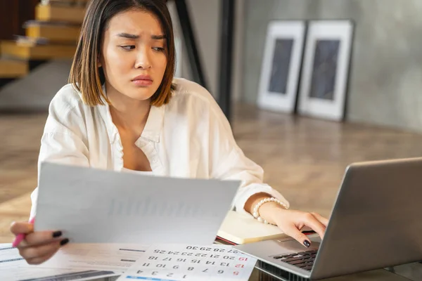 Сумна азіатська молода жінка працює з паперами та ноутбуком. — стокове фото