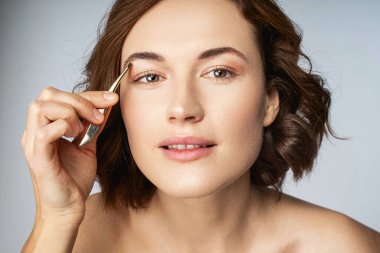 Gülümseyen genç kadın kaş şekillendirme için maşa kullanıyor.