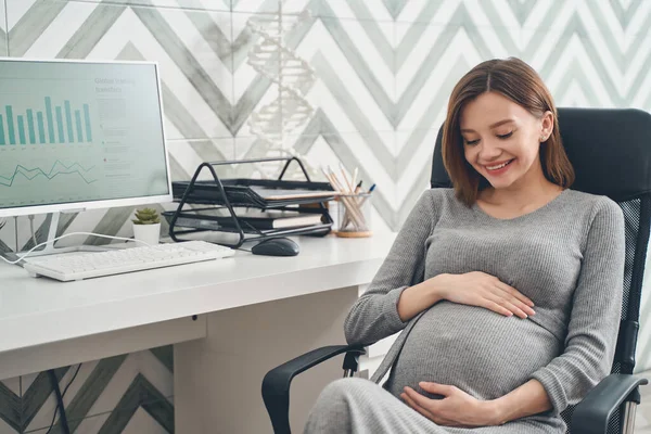 Glädjande gravid dam sitter vid bordet med dator på kontoret — Stockfoto