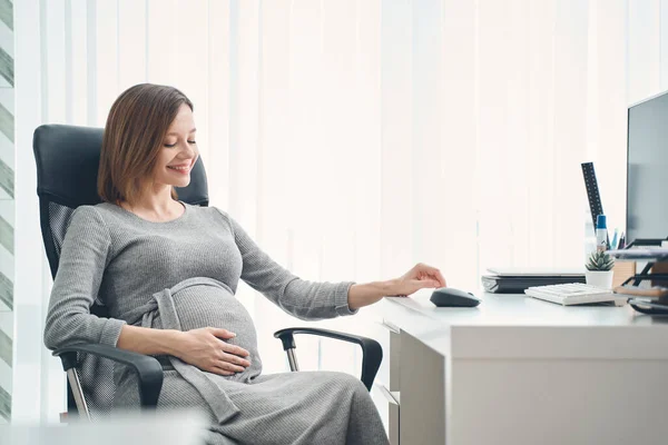 Cute expectant woman sitting at office desk and smiling — Stock Photo, Image