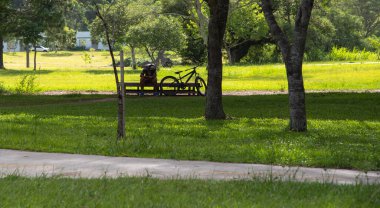 Parkın gölgesinde oturan genç bir adam cep telefonuyla oynuyor. Yanınızdaki bisiklet, sizin ulaşım aracınız. Şehir manzarası ve modern dünyanın eğilimi. Spor ve teknoloji..