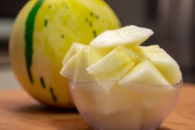 Tropikal meyve parçaları küçük bir kasede insan tüketimi için servis edilen parça parça salatalıklı melo. Sahnenin arka planında yeşil çizgili melez çeşitliliğinde sarı bir kavun meyvesi var..