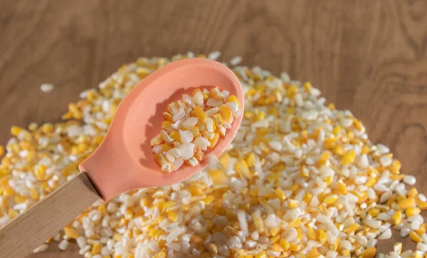 A wooden spoon with a silicone tip filled with mixed yellow and white hominy corn. Below and on a wooden table are many seeds of canjiquinha corn.