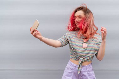 Güneş gözlüklü güzel, gülümseyen hippi kadının moda portresi ve renkli gri duvara karşı akıllı telefonlu renkli kızıl saçlar.