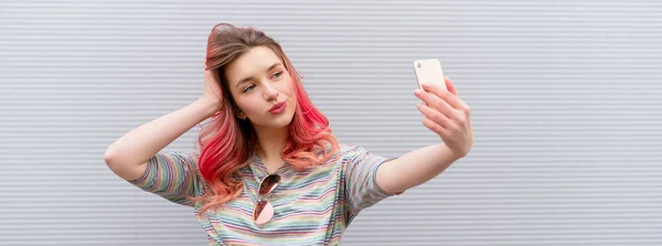 Piękna kobieta z kolorowymi różowymi włosami, bezprzewodowymi słuchawkami i smartfonem na szarym tle — Zdjęcie stockowe