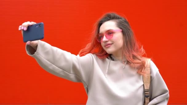 선글라스를 쓰고 빨간 머리를 하고 밝은 오렌지색 벽에 스마트폰을 갖고 있는 예쁘게 웃고 있는 이발소 여성의 패션 사진 — 비디오
