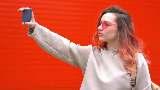 Модный портрет красивой улыбающейся хипстерши в солнцезащитных очках и цветных рыжих волосах со смартфоном у красочной оранжевой стены — стоковое видео