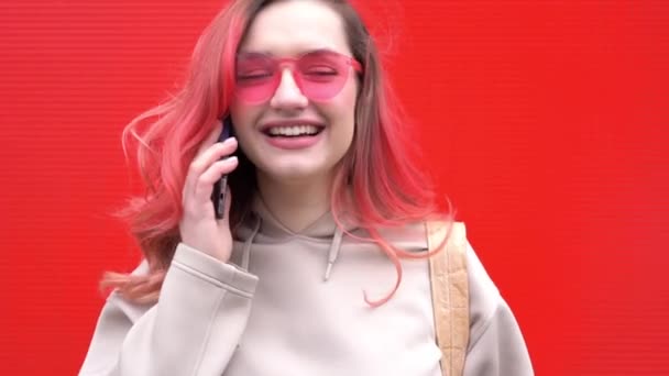 Modeporträt einer hübsch lächelnden Hipsterfrau mit Sonnenbrille und gefärbten roten Haaren mit Smartphone vor der orangefarbenen Wand — Stockvideo