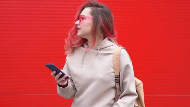 Retrato de moda de mulher hipster muito sorridente em óculos de sol e cabelos vermelhos coloridos com smartphone contra a parede laranja colorida — Vídeo de Stock