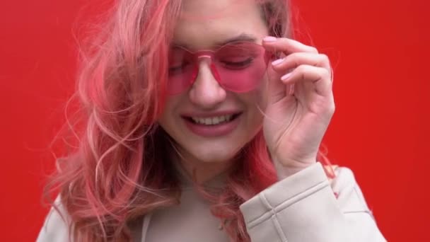 Retrato de moda de mujer hipster bastante sonriente en gafas de sol y pelos rojos de colores con teléfono inteligente contra la colorida pared naranja — Vídeos de Stock