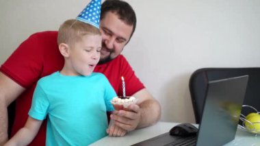 Babasıyla birlikte küçük bir çocuk doğum gününü internette akrabaları ve arkadaşlarıyla deftere bakarak kutluyor. Mumu üfle.