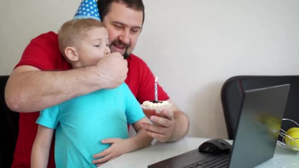 Маленький мальчик с отцом празднуют день рождения онлайн, разговаривая по блокноту с родственниками и друзьями. Задуй свечу. — стоковое видео