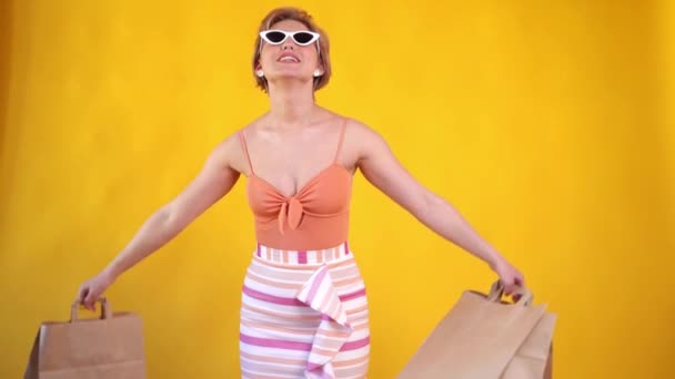 Divat szépség hűvös fiatal lány bevásárló táskák nyári ruha és napszemüveg felett színes narancs háttér — Stock videók