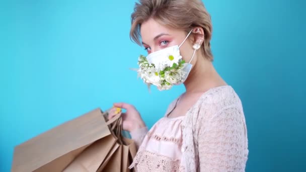 Moda belleza fresca chica joven en máscara médica diseñada con flores naturales de pie con bolsas de compras en vestido de verano sobre la pared azul — Vídeos de Stock