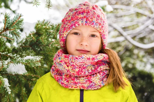 Χειμώνα πορτρέτο του ένα χαριτωμένο κοριτσάκι κάτω από τις χιονοπτώσεις φοράει πράσινο σακάκι, πολύχρωμα πλεκτά καπέλο και κασκόλ — Φωτογραφία Αρχείου