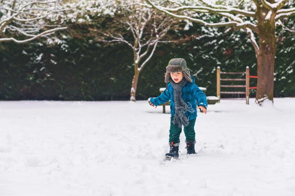 Söpö poika leikkimässä talvipuistossa. Lapsi pitää hauskaa ulkona, juoksee lumella, yllään lämmin sininen takki, hattu ja huivi — kuvapankkivalokuva
