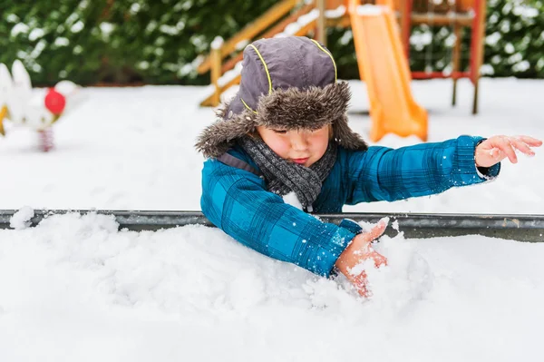 Menino adorável brincando em caixa de areia cheia de neve, usando chapéu quente — Fotografia de Stock